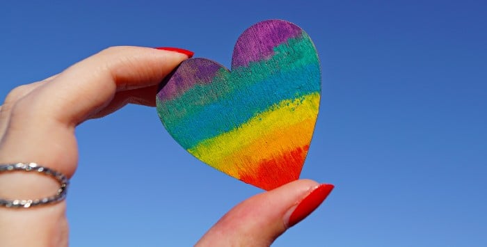 Hand holding a heart painted with the colors of the LGBTQ flag